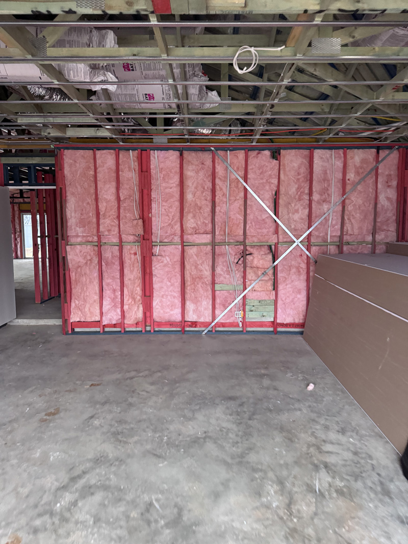 Pink wall batts in a steel-framed wall, with plasterboard stacked alongside