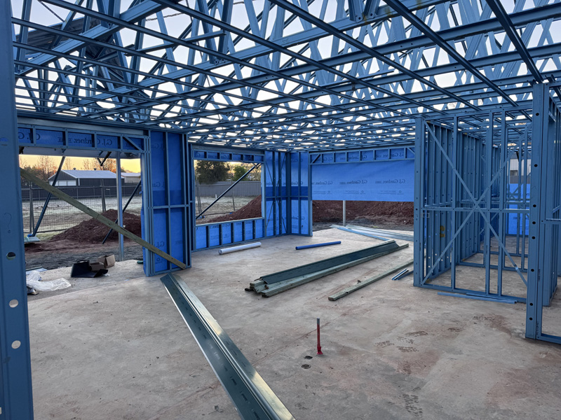 Inside a blue steel house frame, with roof trusses overhead and steel lengths on the slab