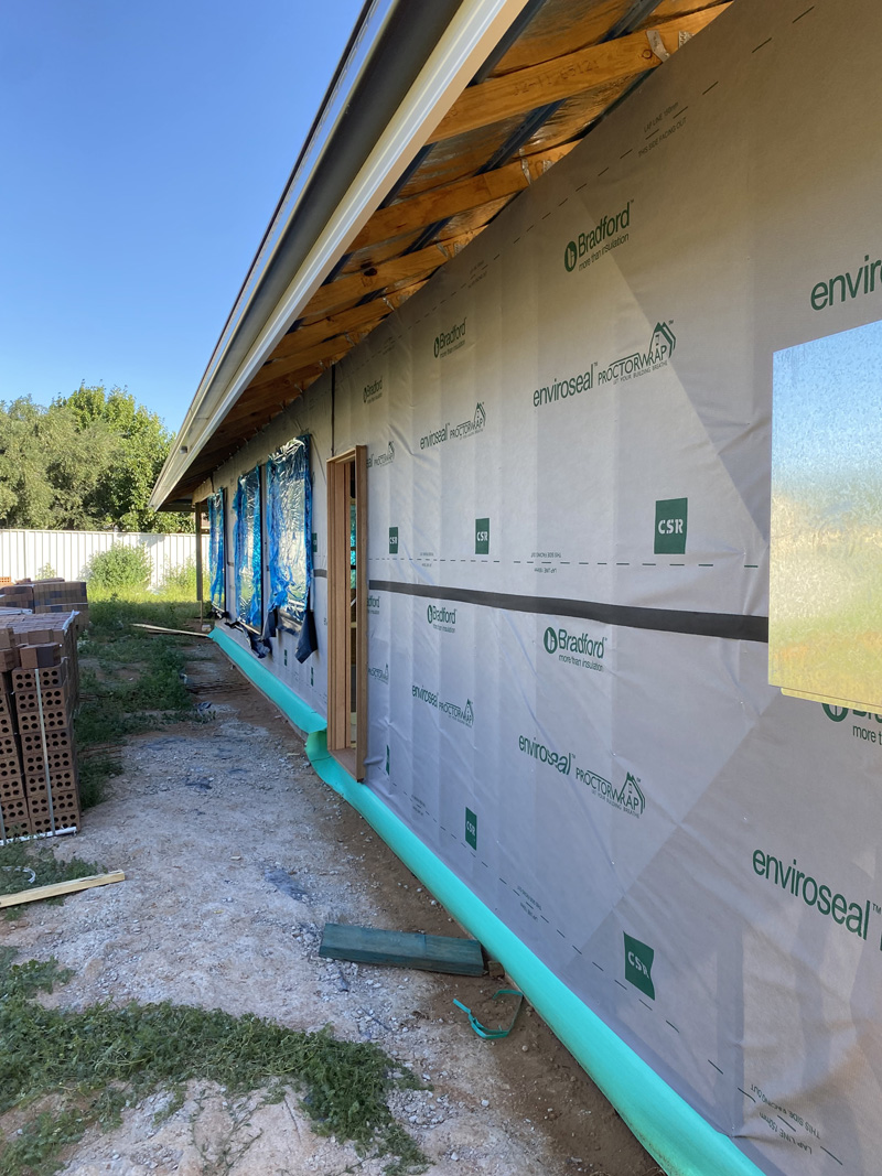 Wall-wrapped new house beside a gravel side path, with bricks stacked nearby